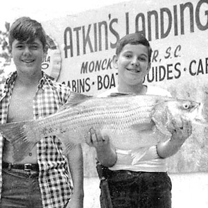 Two boys standing in front of Atkin's Landing sign proudly hold a large fish